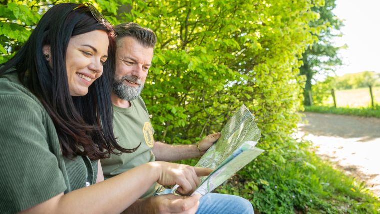 A couple is sitting relaxed under trees and looking at a map. They seem to be enjoying their surroundings and planning their trip.