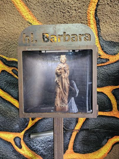 Een standbeeld van de heilige Barbara in een glazen container. De achtergrond toont de kleurrijke muurschildering van de Ulmener Maar-stollen.