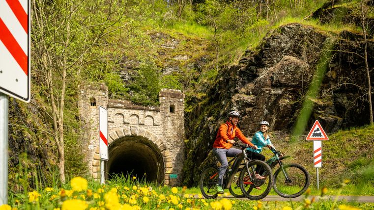 Cyclists enjoy nature in front of an old tunnel. Surrounded by green meadows and blooming dandelions.