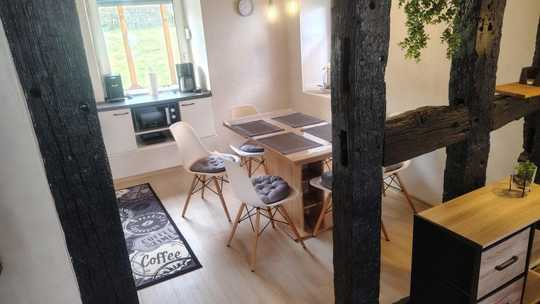 A bright kitchen with a modern dining table and chairs. The room has rustic wooden beams and large windows that provide plenty of daylight.