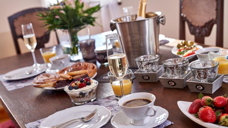 Ein schön gedeckter Tisch mit frischem Frühstück, darunter Croissants, Joghurt mit Beeren und frischen Erdbeeren. Gläser mit Champagner und Orangensaft runden das Bild ab.