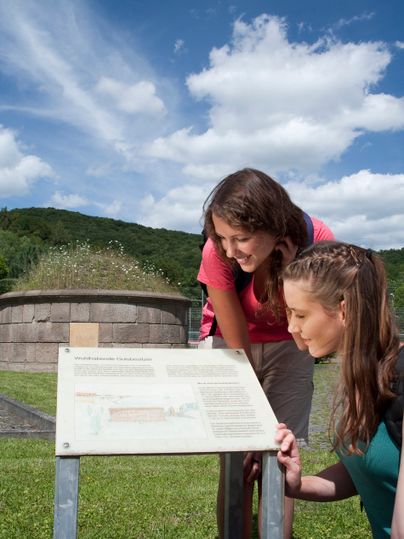 Zwei Frauen lesen eine Informationstafel im Freien vor einem runden Steindenkmal.