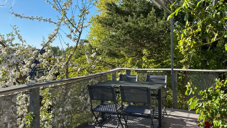 A cozy balcony with a table and chairs, surrounded by blossoming trees. The sun is shining and the sky is clear.