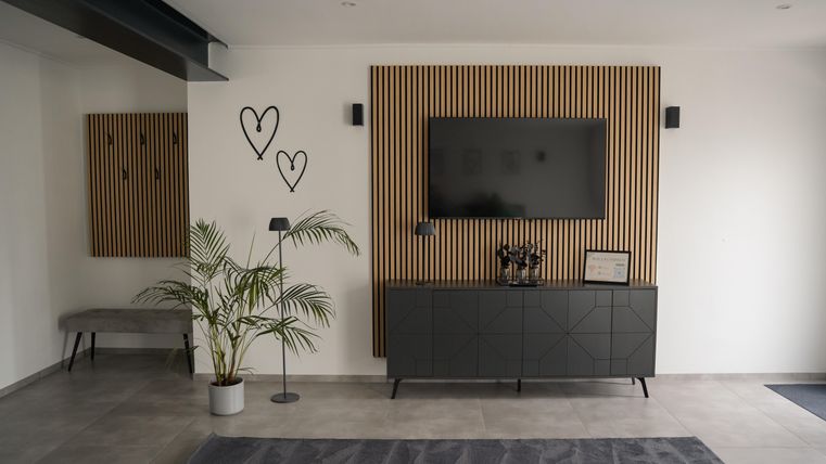 Een moderne woonkamer met houten lambrisering en een televisie aan de muur. Een plant en een zwart dressoir maken het eenvoudige, elegante interieur compleet.
