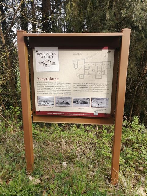 An information sign about an archaeological excavation with a plan and images. The sign is surrounded by trees and greenery.