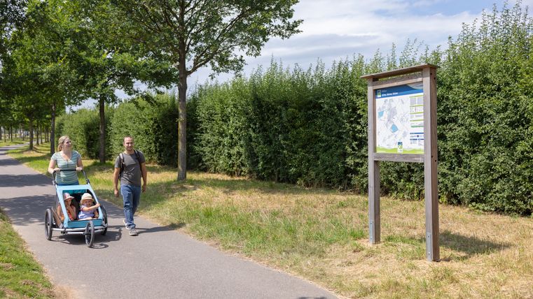 A couple walks with a baby carriage on a paved path next to an information board in the countryside.