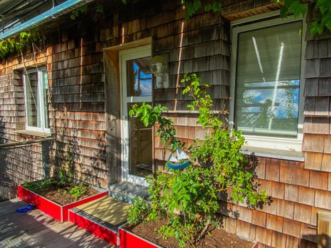 Een met hout bekleed huis met een ingang en twee ramen. Voor het huis staan rode plantenkasten met groene planten.