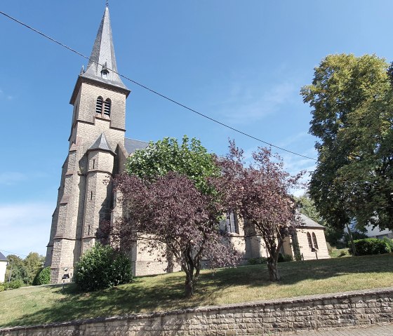De parochiekerk van St Bartholomew met zijn hoge, spitse toren staat omringd door bomen onder een helderblauwe hemel., © TI Bitburger Land - Steffi Wagner