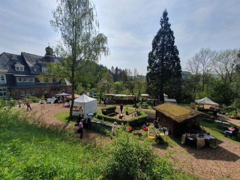 Een mooie tuin met stands, verkopers en bezoekers. Op de achtergrond staat een groot gebouw omringd door bomen en groen.