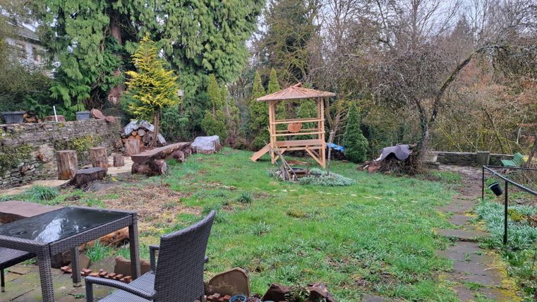 A garden with green grass and several trees. In the background, there is a wooden pergola, and a table with chairs is visible in the foreground.