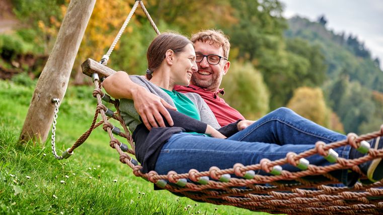 A couple lies smiling in a hammock on a meadow surrounded by trees.