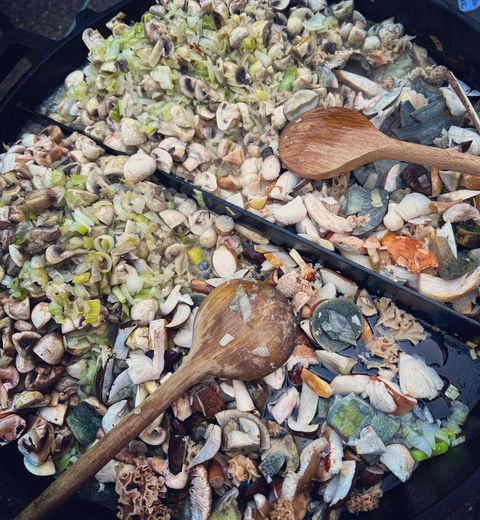 A large pan with a mixture of fresh seafood, vegetables, and herbs. Two wooden spoons are ready for stirring.