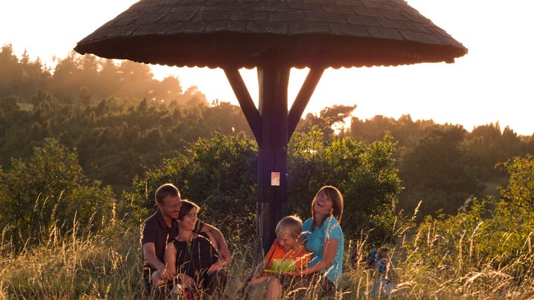 Family sitting under a wooden shelter in a meadow at sunset.