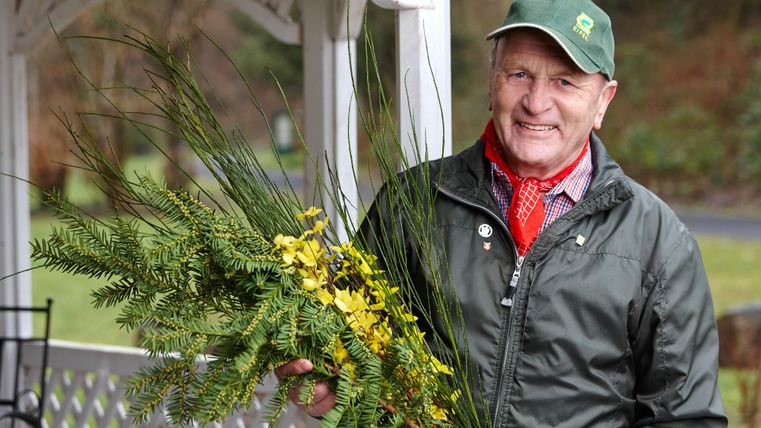 Een oudere man draagt een krans van groene takken en gele bloemen. Hij staat onder een overdekte ruimte in een park.