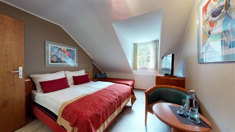 Ein gemütliches Zimmer mit einem Doppelbett, roten Kissen und moderner Dekoration. Die großen Fenster lassen viel Tageslicht herein und bieten einen einladenden Blick.