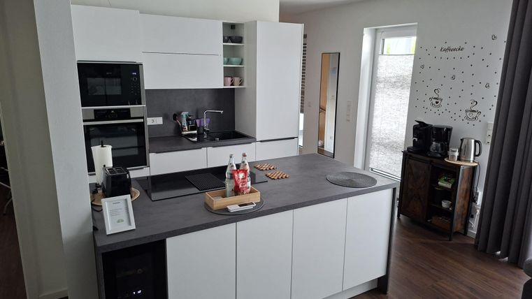Eine moderne Küche mit weißen Möbeln und einer großen Kochinsel. Es gibt Geräte wie einen Ofen und einen Kühlschrank, sowie eine helle Fensterfront.