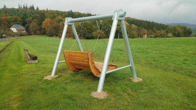 Eine große Holzschaukel steht auf einer grünen Wiese. Im Hintergrund sind Bäume und ein paar Häuser sichtbar.