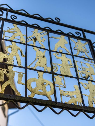 An artistic metal frame with golden figures representing various activities. The background is a clear blue sky.