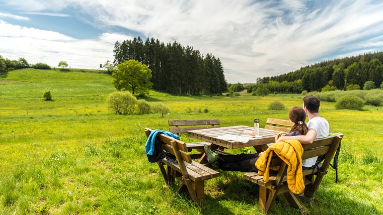 Un couple est assis sur un banc à une table en bois dans un paysage verdoyant avec des arbres en arrière-plan.