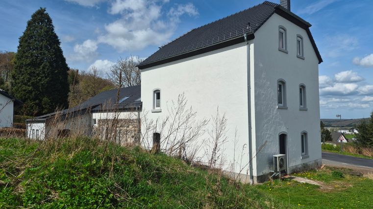 Een modern, wit huis staat op een heuvel in een landelijke omgeving. Op de achtergrond zijn bomen en een blauwe lucht te zien.