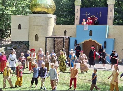 Eine lebhafte Szenerie mit vielen Kindern und Erwachsenen in bunten Kostümen. Im Hintergrund ist ein großes, fantasievolles Gebäude mit einer goldenen Kuppel zu sehen.