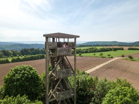 Holzaussichtsturm in ländlicher Landschaft mit Feldern und Wäldern im Hintergrund.