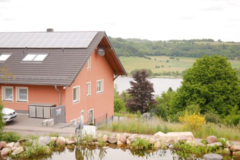 De afbeelding toont een modern huis met een zonnepaneel op het dak. Op de achtergrond strekt zich een groene landschap uit met een meer.