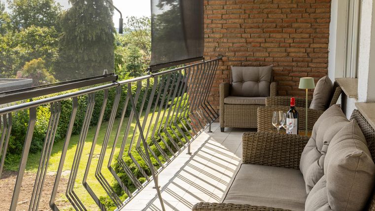 Een gezellige terras met een rattanbank en een fauteuil. Op de tafel staan een fles wijn en glazen, omgeven door een groene uitzicht.