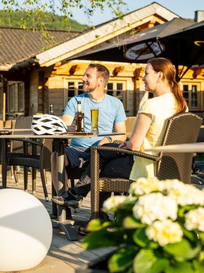 Zwei Personen sitzen an einem Tisch im Freien, mit Getränken und einem Fahrradhelm auf dem Tisch.