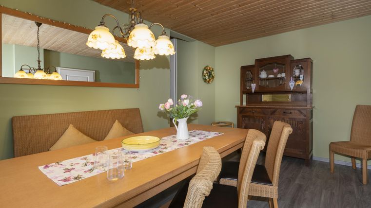 Een gezellige eettafel van hout in een lichte kamer. De muren zijn groen geschilderd en een lamp met meerdere lampen hangt aan het plafond.