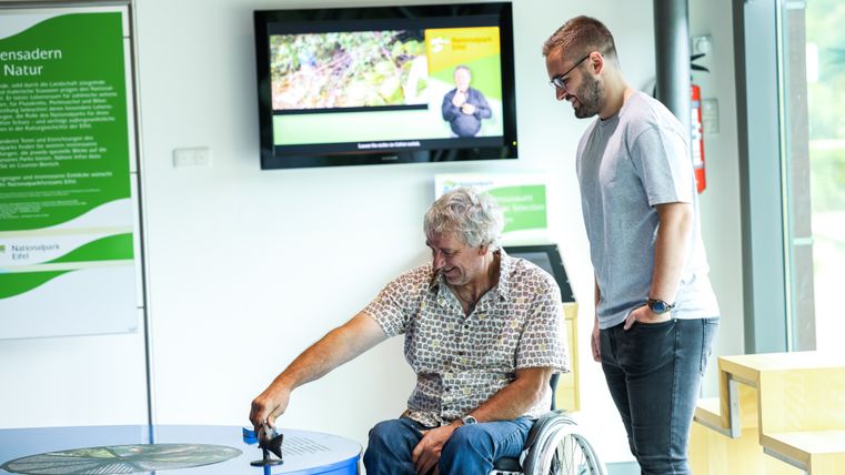 Zwei Personen sind in der barrierefreien Ausstellung im Nationalpark Tor Rurberg. Eine von den beiden Sitzt im Rollstuhl und interagiert mit den barrierefreien Modellen der Ausstellung.
