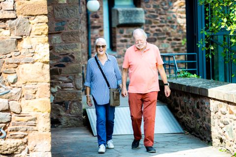 Ein älteres Paar geht nebeneinander auf einem Weg aus Stein. Die beiden sind auf der Burg Hengebach. Um sie herum sind hohe Mauern aus braunem Stein. Es ist ein sonniger Tag.