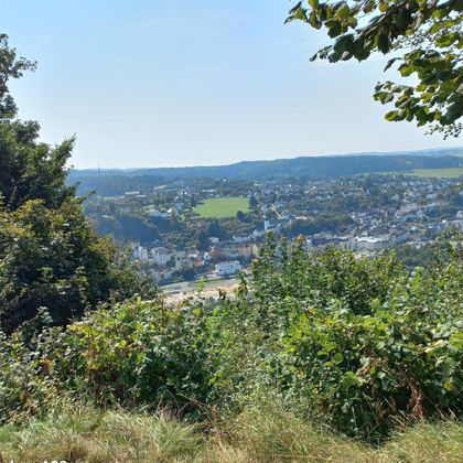 Een landschap met uitzicht op een stad, omgeven door groene heuvels. De lucht is helder en blauw.