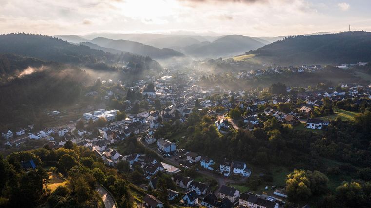 Eine malerische Landschaft mit einem kleinen Dorf in einem Tal. Sanfte Hügel und Nebel ergänzen die ruhige Atmosphäre.
