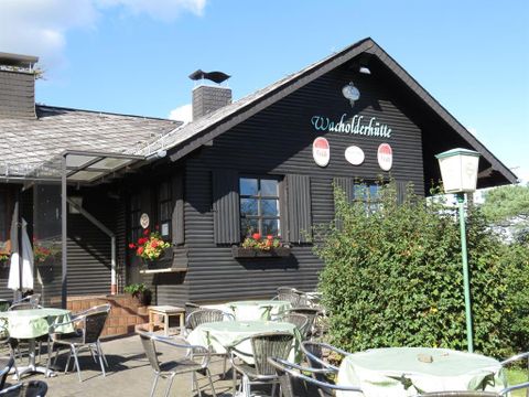A traditional black cottage named "Waldschlötte" stands in the foreground. Tables and chairs are outdoors, surrounded by green plants and flowers.