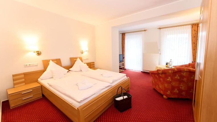 Een gezellige hotelkamer met een tweepersoonsbed en een rode tapijt. Er is een zithoek met een bank en veel daglicht.
