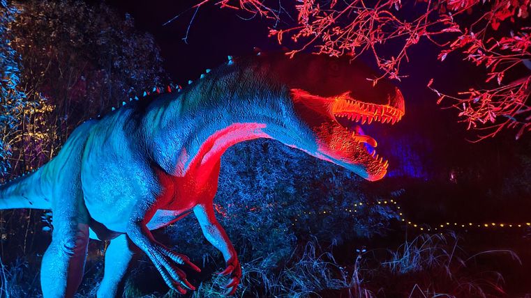 Un dinosaure lumineux se tient dans un environnement sombre, entouré de lumières colorées. Les couleurs sont intenses et donnent à la scène une atmosphère excitante.