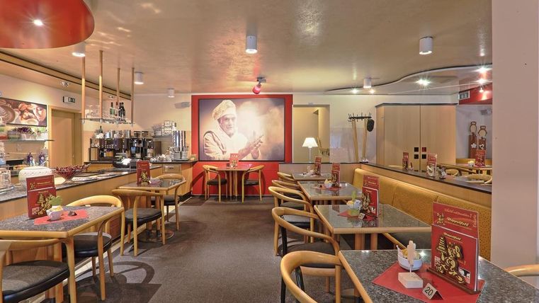 Cozy café with wooden furniture, tables, and chairs. On the wall, a large picture of a baker. Red accents and menus on the tables.