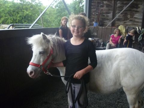 Au premier plan de l'image se trouve une fille en t-shirt noir et pantalon gris devant un poney blanc. Dans sa main gauche, elle tient une corde noire, tandis que de sa main droite, elle gratte le poney sous la tête. Le poney porte un harnachement rouge. À l'arrière-plan, on peut voir quelques personnes. Elles se trouvent dans une écurie.
