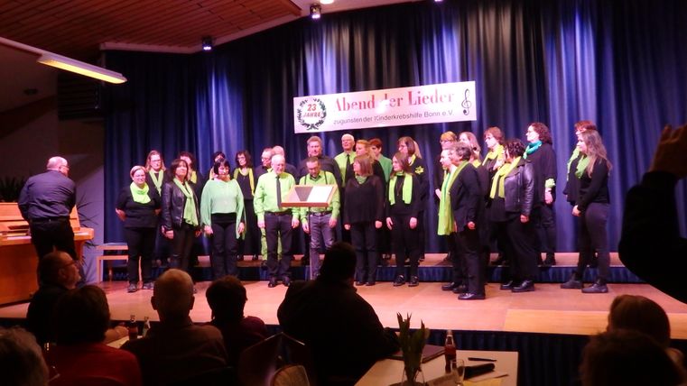 A choir in colorful outfits sings on a stage. In the audience, many listeners are enjoying the performance.