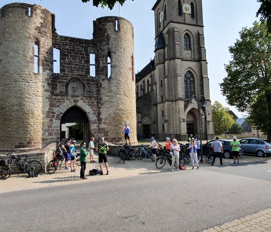 Groupe de cyclistes devant une porte historique et une église, entourés d'arbres et de voitures garées., © Tourist-Information Bitburger Land