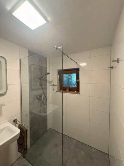 A modern bathroom with a glass shower and a small window. The walls are white and the floor is gray.