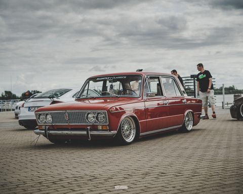 Ein roter Oldtimer steht auf einem Parkplatz. Im Hintergrund sind weitere Autos und Personen zu sehen.