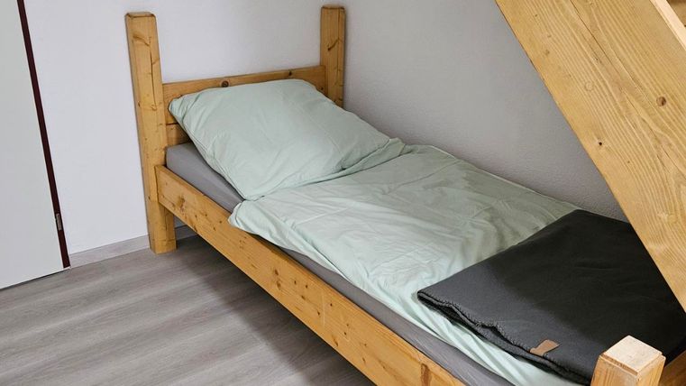 A simple wooden bed stands in a room with bright walls. Next to the bed, straight steps are visible.