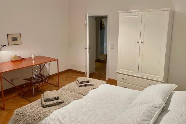 Ein modernes Schlafzimmer mit einem Doppelbett und einem weißen Kleiderschrank. Ein Schreibtisch mit einem Stuhl ist im Hintergrund sichtbar.