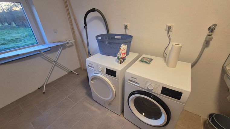 Une salle de lavage avec une machine à laver et un sèche-linge. À l'arrière-plan se trouvent un panier à linge et une table à repasser.