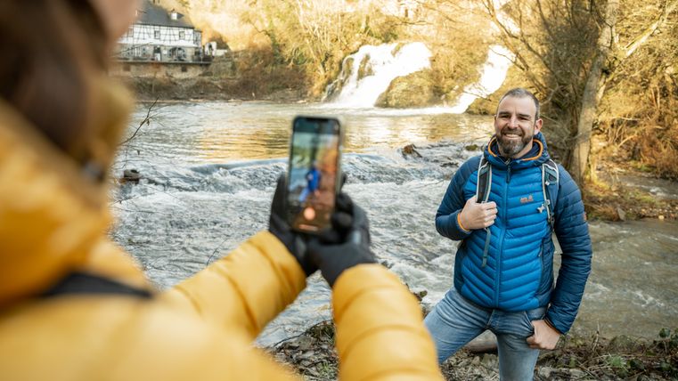A man in a blue jacket poses smiling for a photo by the river while another person takes a photo of him with a smartphone.