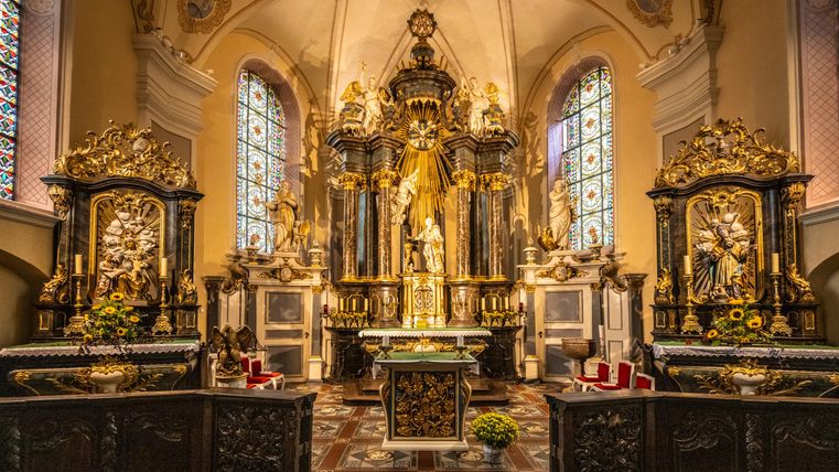 A beautiful church altar with elaborate carvings and golden accents. Colorful windows let the light shine impressively inside.