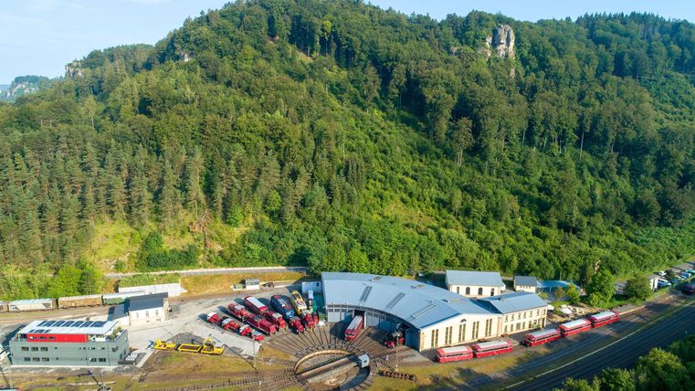 An impressive landscape with a modern train depot and numerous trains. In the background, green hills and trees can be seen.