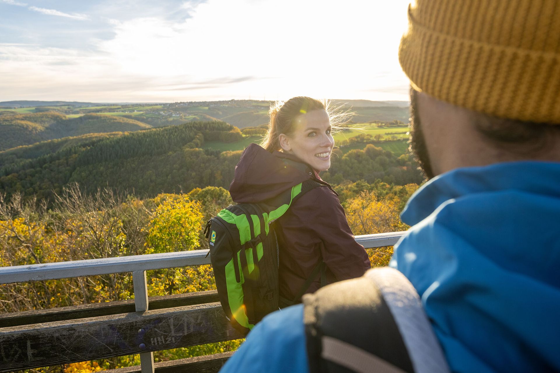 Eine Frau dreht sich Richtung Kamera, dahinter geht die Sonne über Wald unter